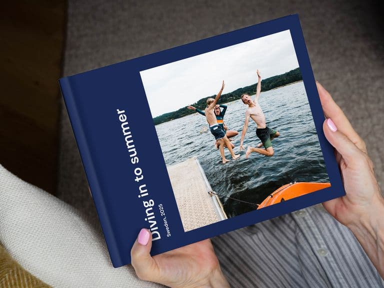 Person holding a photo book titled "Diving in to summer," featuring an image of two people jumping into a lake from a dock.