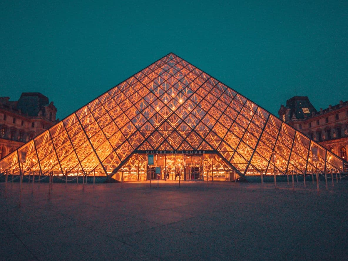 Illuminated glass pyramid entrance of the Louvre Museum at night, surrounded by historic buildings under a dark turquoise sky.