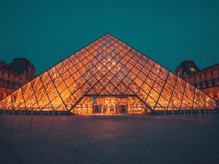 Illuminated glass pyramid entrance of the Louvre Museum at night, surrounded by historic buildings under a dark turquoise sky.
