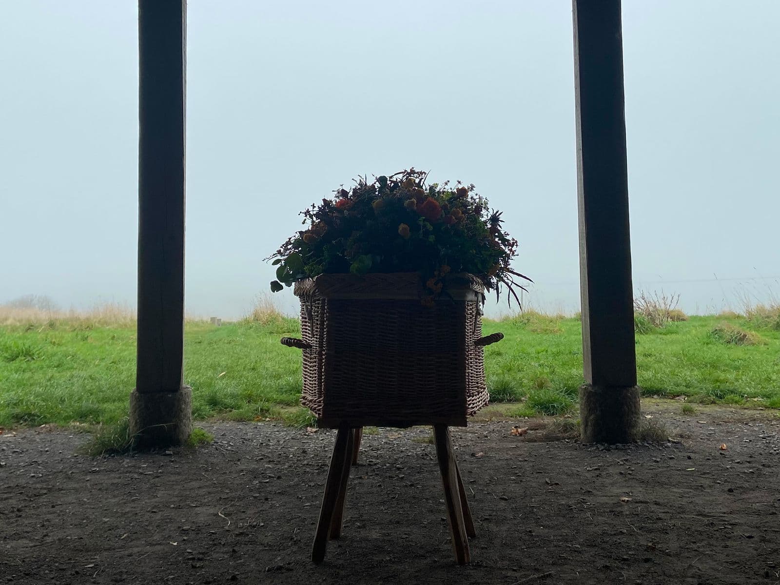 Wicker planter with flowers on a wooden stand centered between two support posts, overlooking a foggy green field.