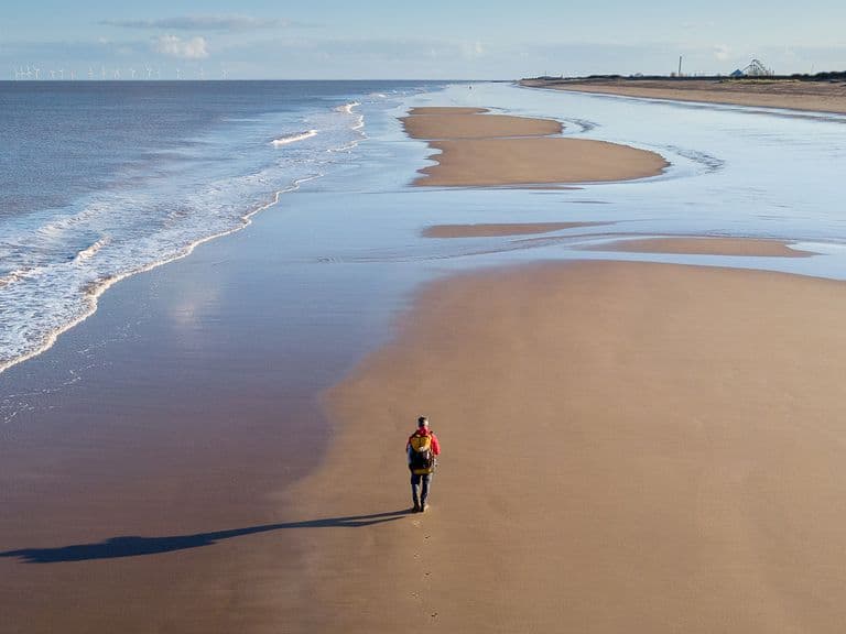 Person walking alone on a vast, empty beach with gentle waves and a clear sky, casting a long shadow on the sand.