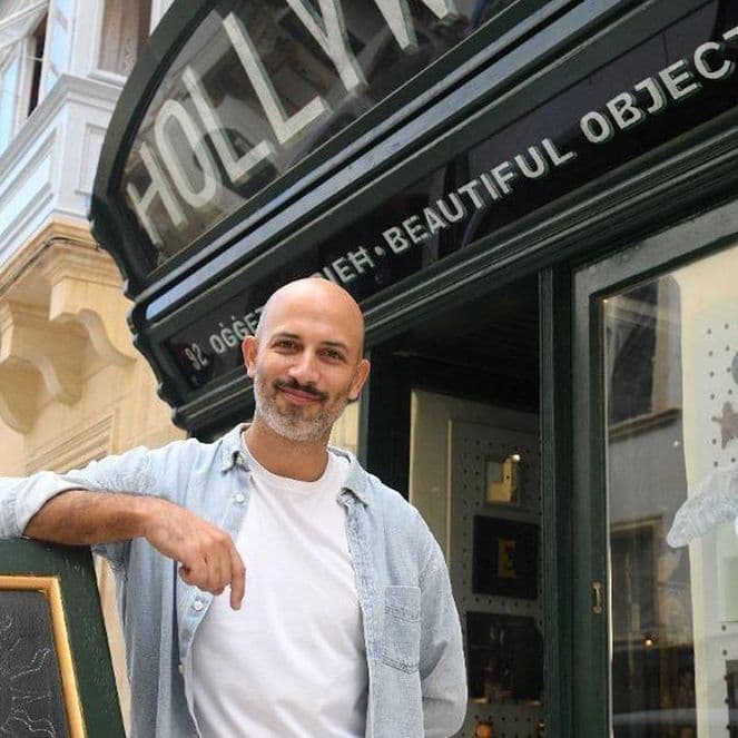 A man stands smiling outside a store named "Hollywood" with a chalkboard sign and display window showing various decorative items.