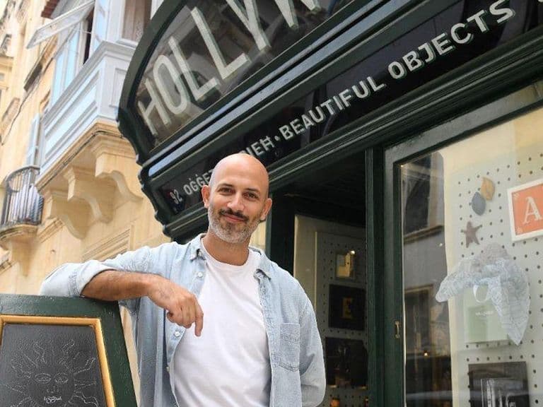 A man stands smiling outside a store named "Hollywood" with a chalkboard sign and display window showing various decorative items.