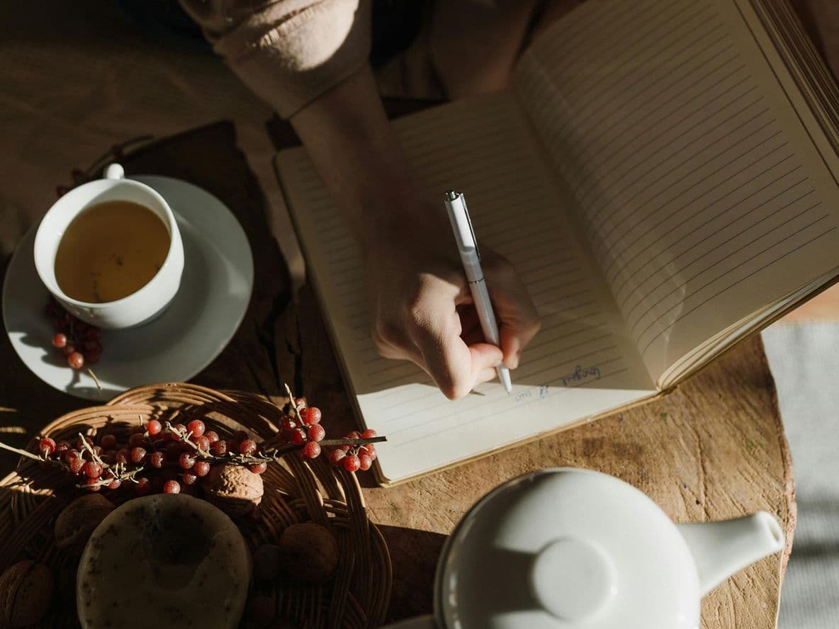 Person writing in a notebook at a table with a cup of tea, a teapot, and a basket of berries in warm, natural light.