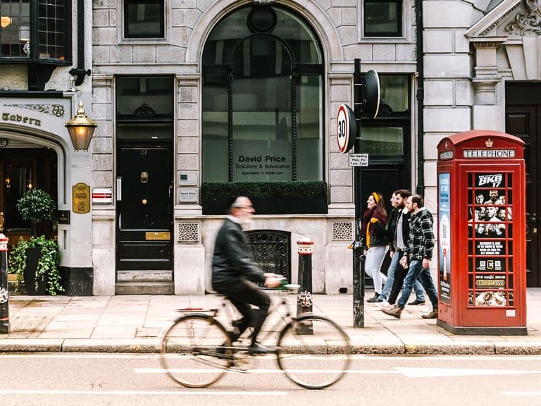 A cyclist passes by a classic red telephone booth and pedestrians on a city street, lined with historic buildings and a pub.