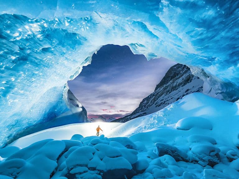 Person in winter clothing stands under a vast, blue ice cave with a view of snowy mountains and a twilight sky in the background.