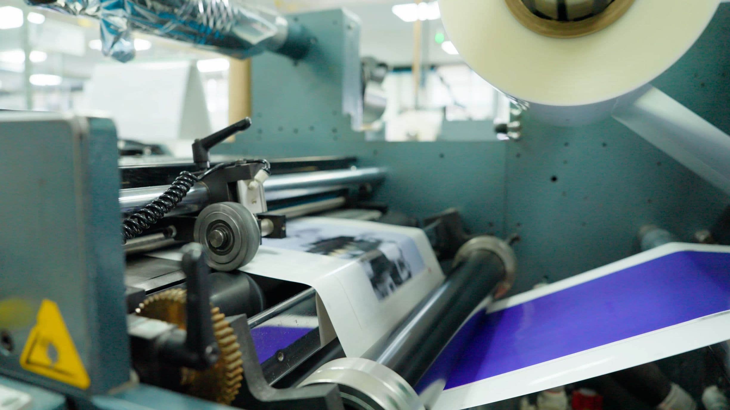 Close-up of a printing press in operation, feeding paper through rollers. The machine is working with blue and white paper.