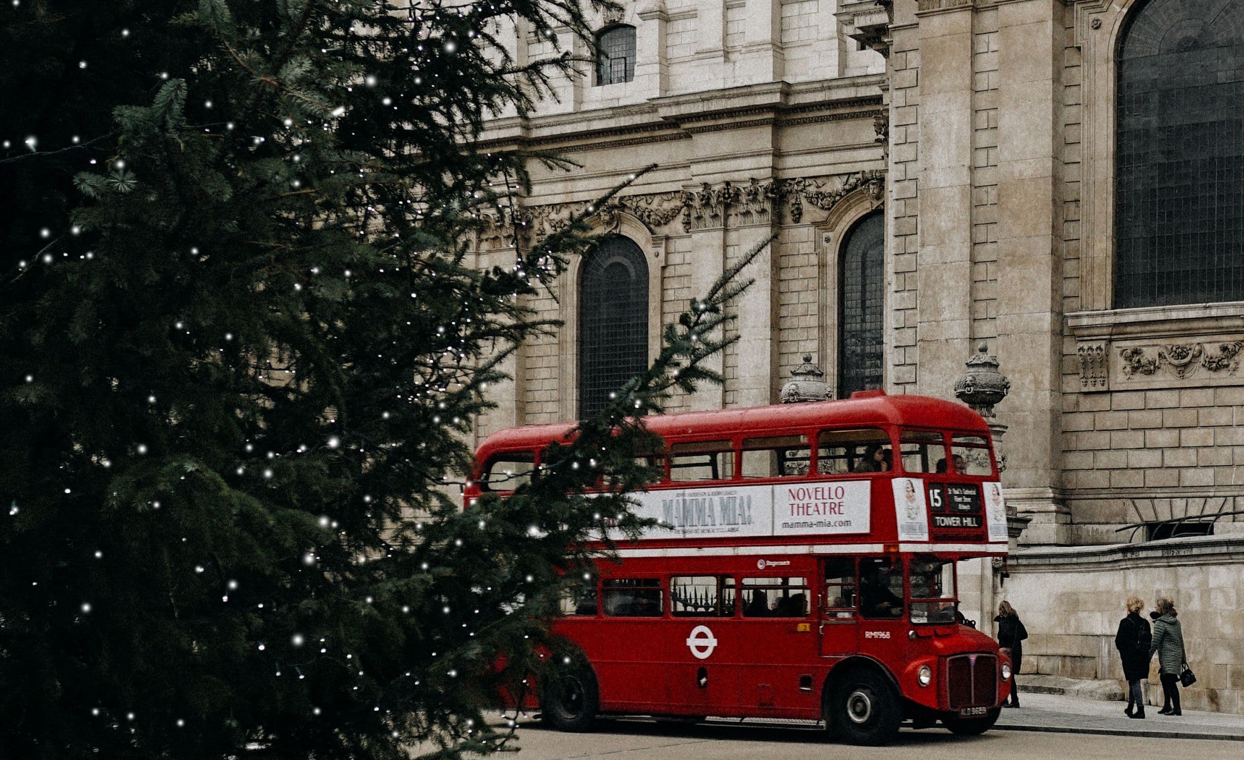 A red double-decker bus passes by a historic stone building, with a large Christmas tree adorned with lights in the foreground.