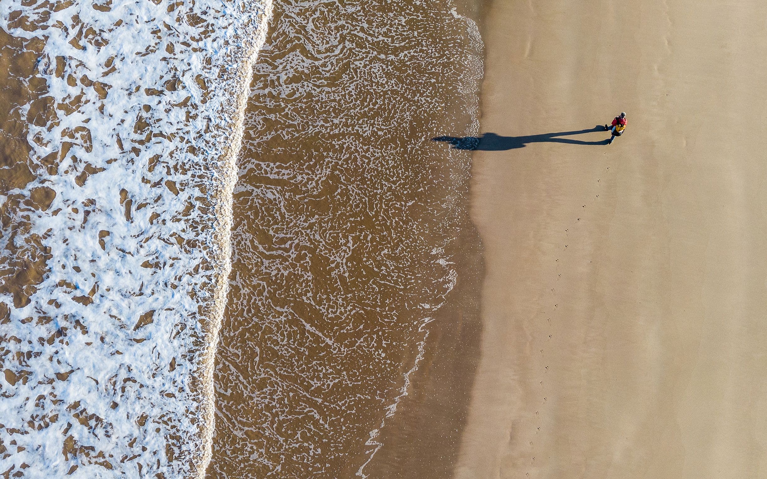Aerial view of a person walking on a sandy beach with gentle waves approaching the shore, casting a long shadow.