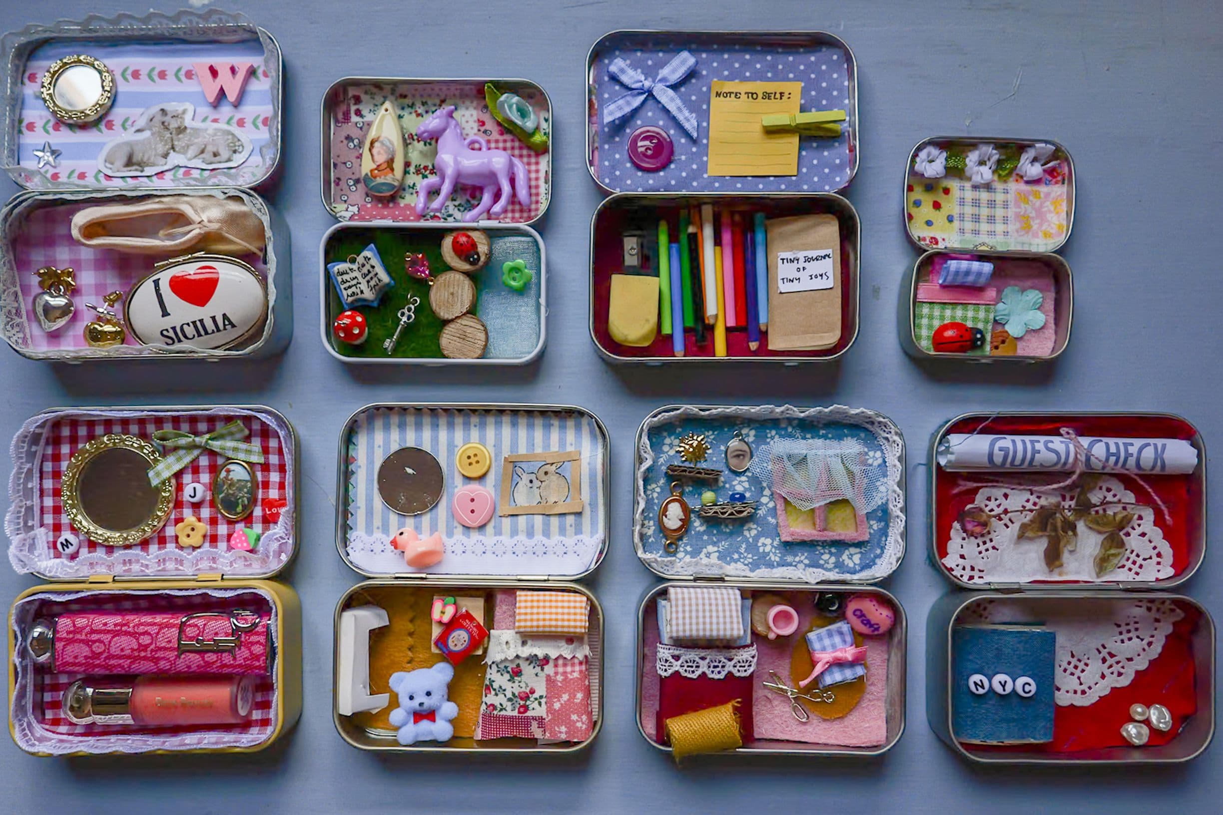 Grid of open small tins, each containing colorful miniature dioramas and objects — buttons, toys, sewing supplies, tiny books and stationery.
