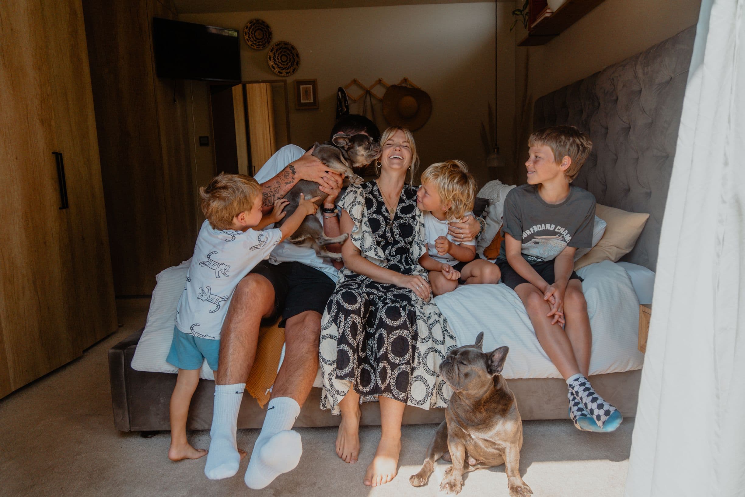 A family of five, including two children, sits on a bed with two dogs, smiling and playing in a warmly lit room.