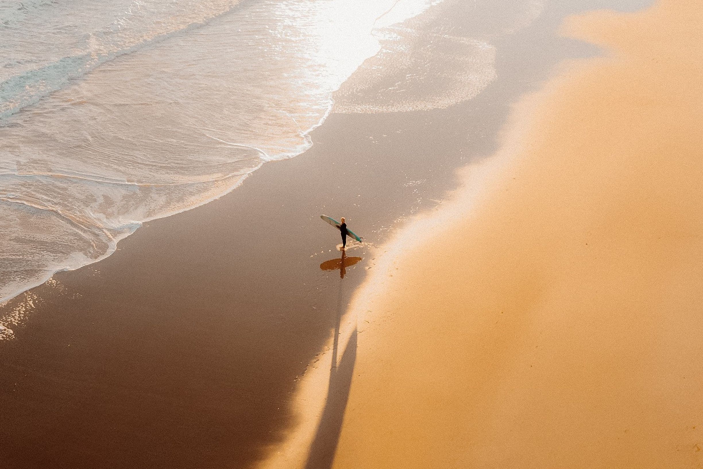 A lone surfer walks along a sunlit, sandy beach, casting a long shadow near the gentle waves.