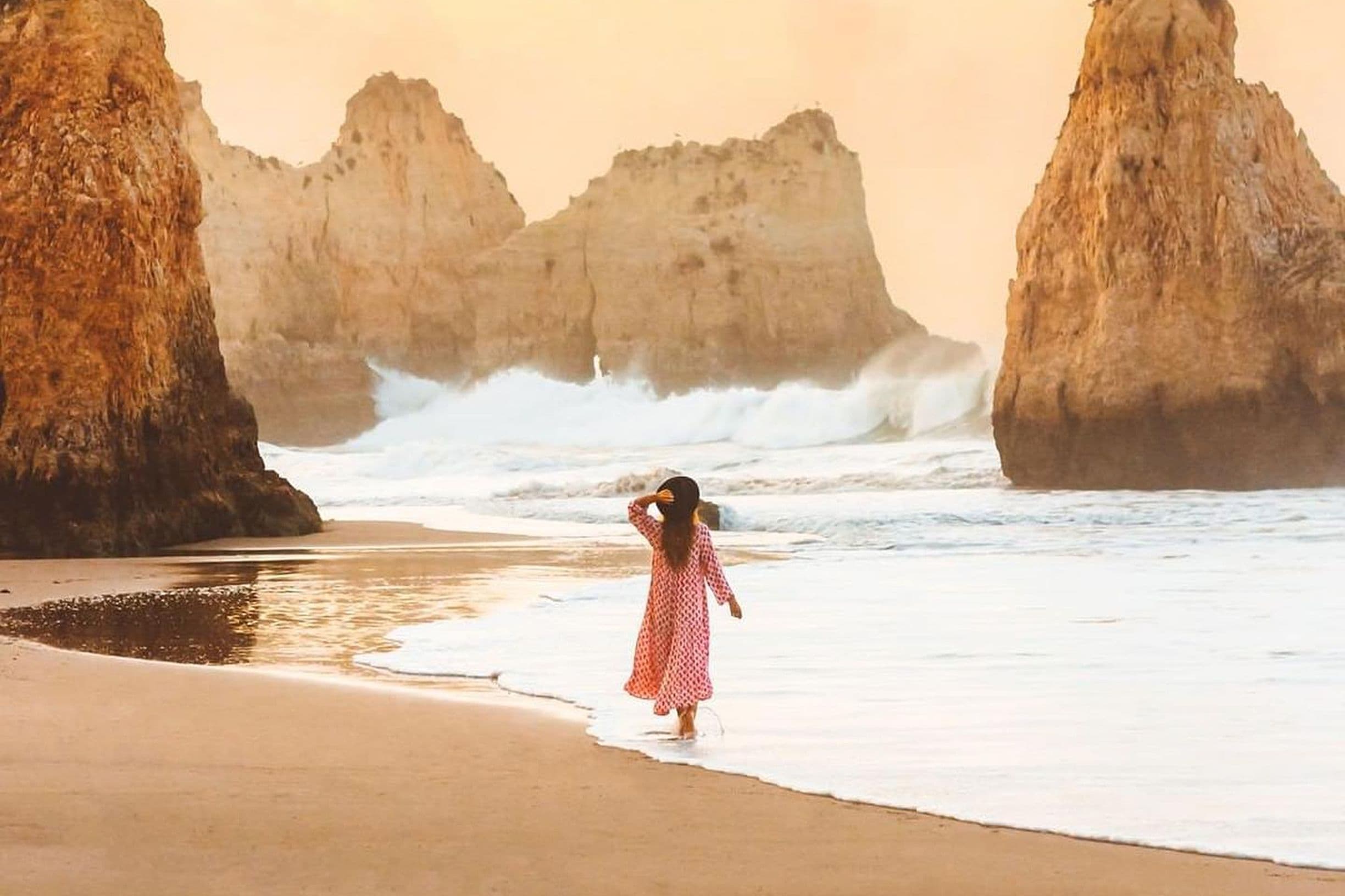 A woman in a red polka dot dress walks along a sandy beach, with large rock formations and waves in the background.