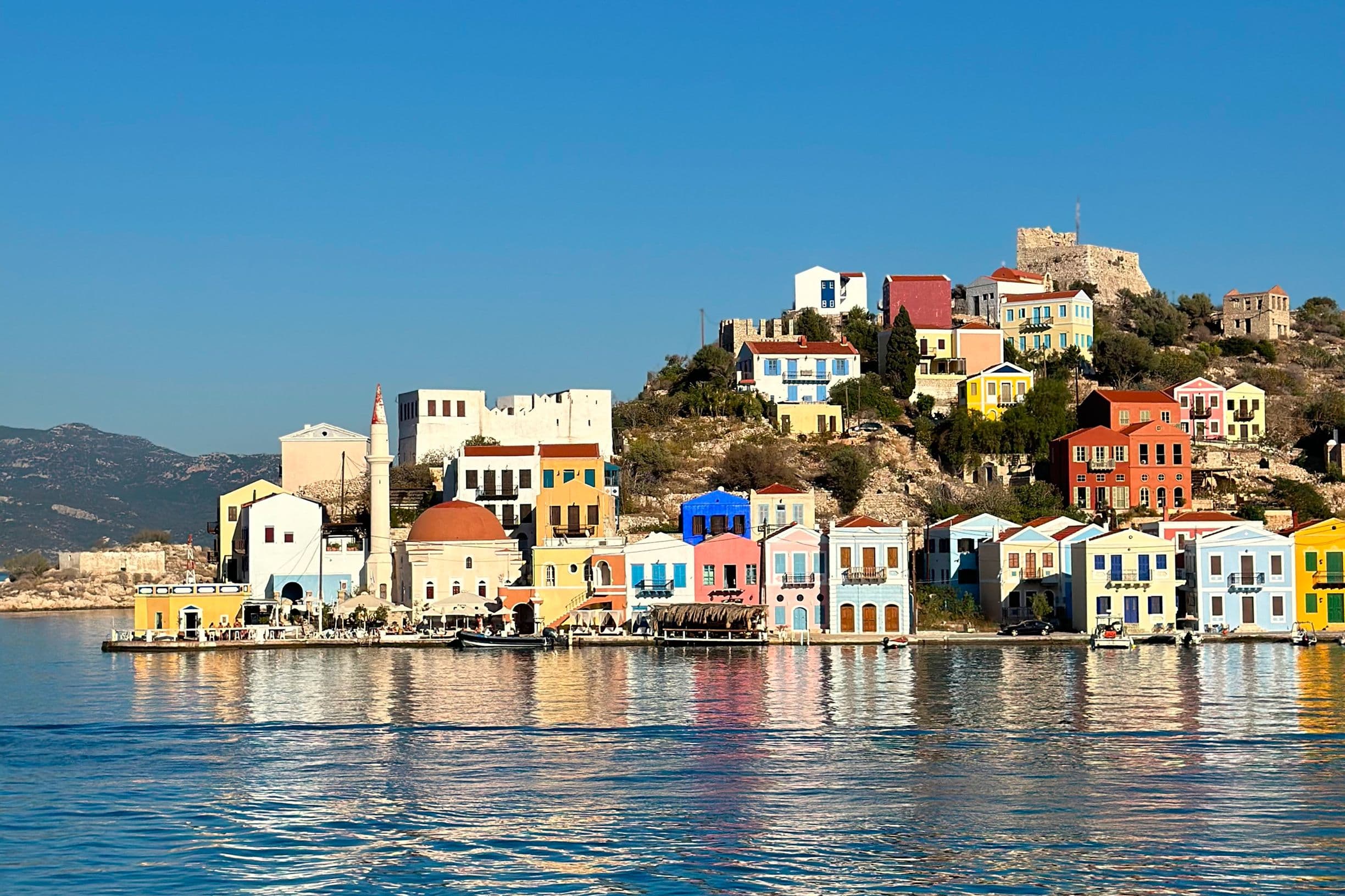 Colorful houses line the waterfront of a picturesque coastal village, reflecting in the calm blue sea under a clear sky.