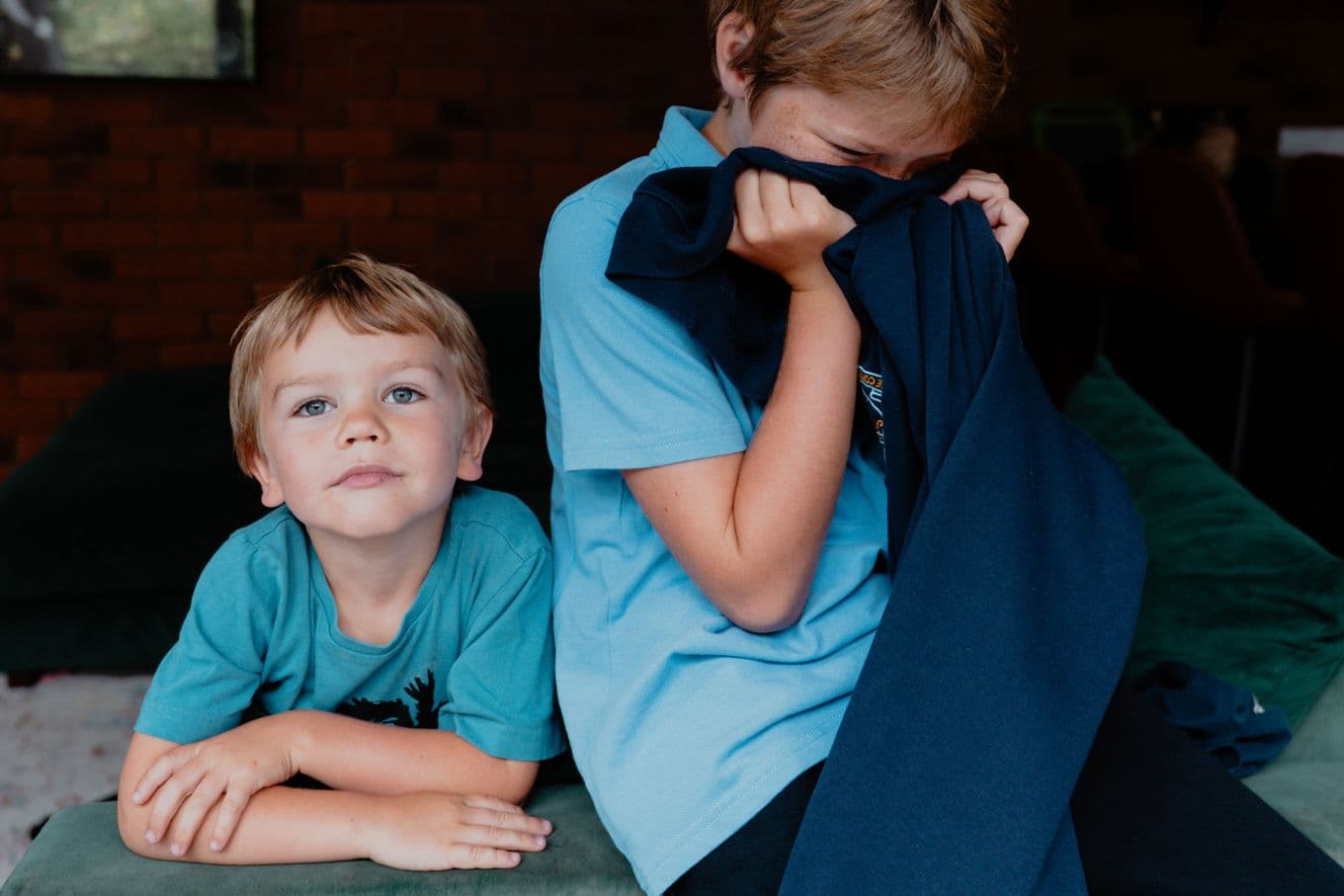 Two young boys in blue shirts; one looks at the camera, while the other hides his face with a dark blue garment.