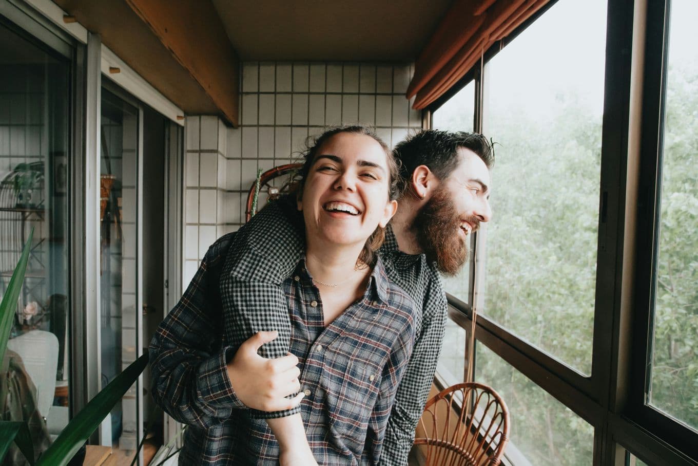 A smiling couple stands on a balcony, with the man playfully leaning on the woman's shoulder. They are surrounded by greenery outside the window.