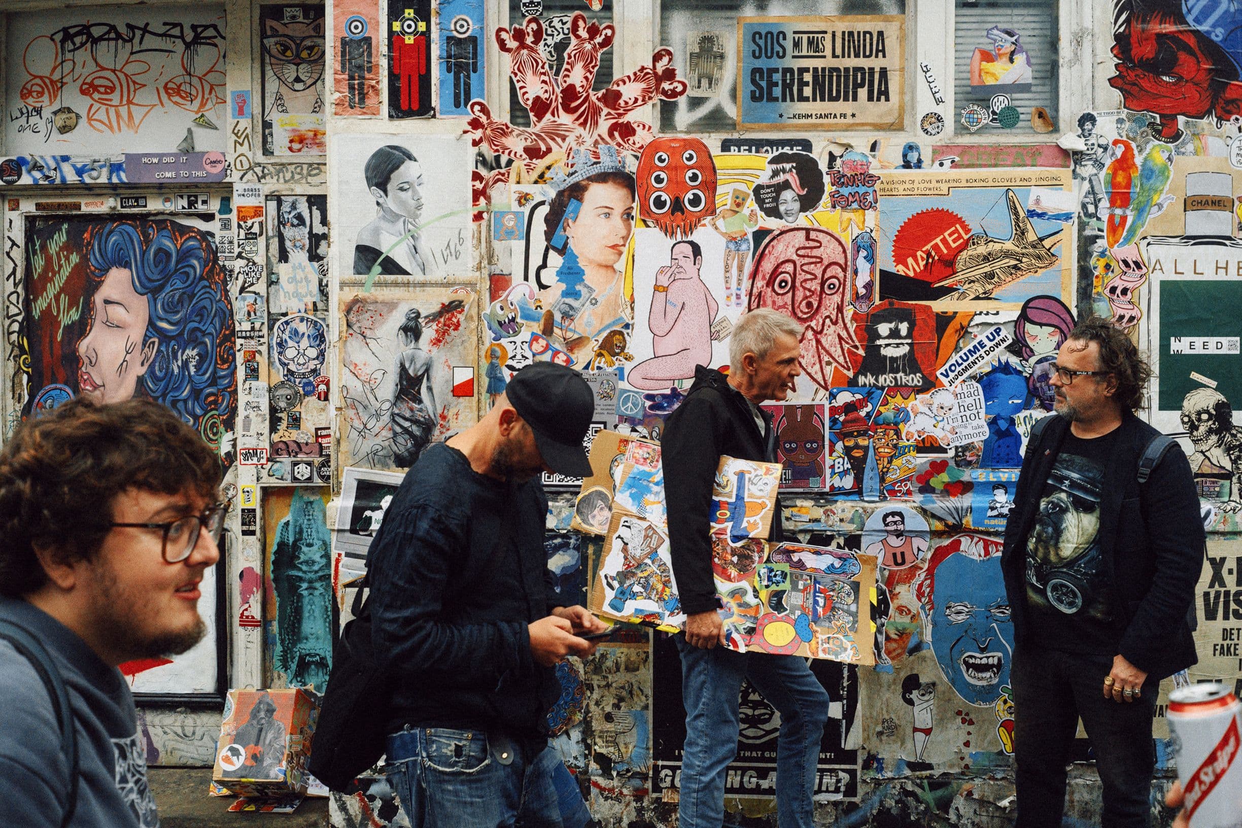 People stand in front of a colorful graffiti-covered wall, featuring various posters and street art.