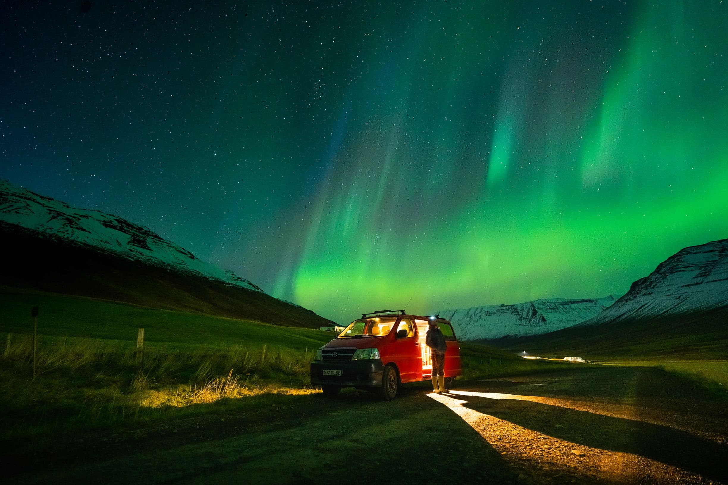 A person stands by a red van under vibrant green auroras in a mountainous landscape at night.