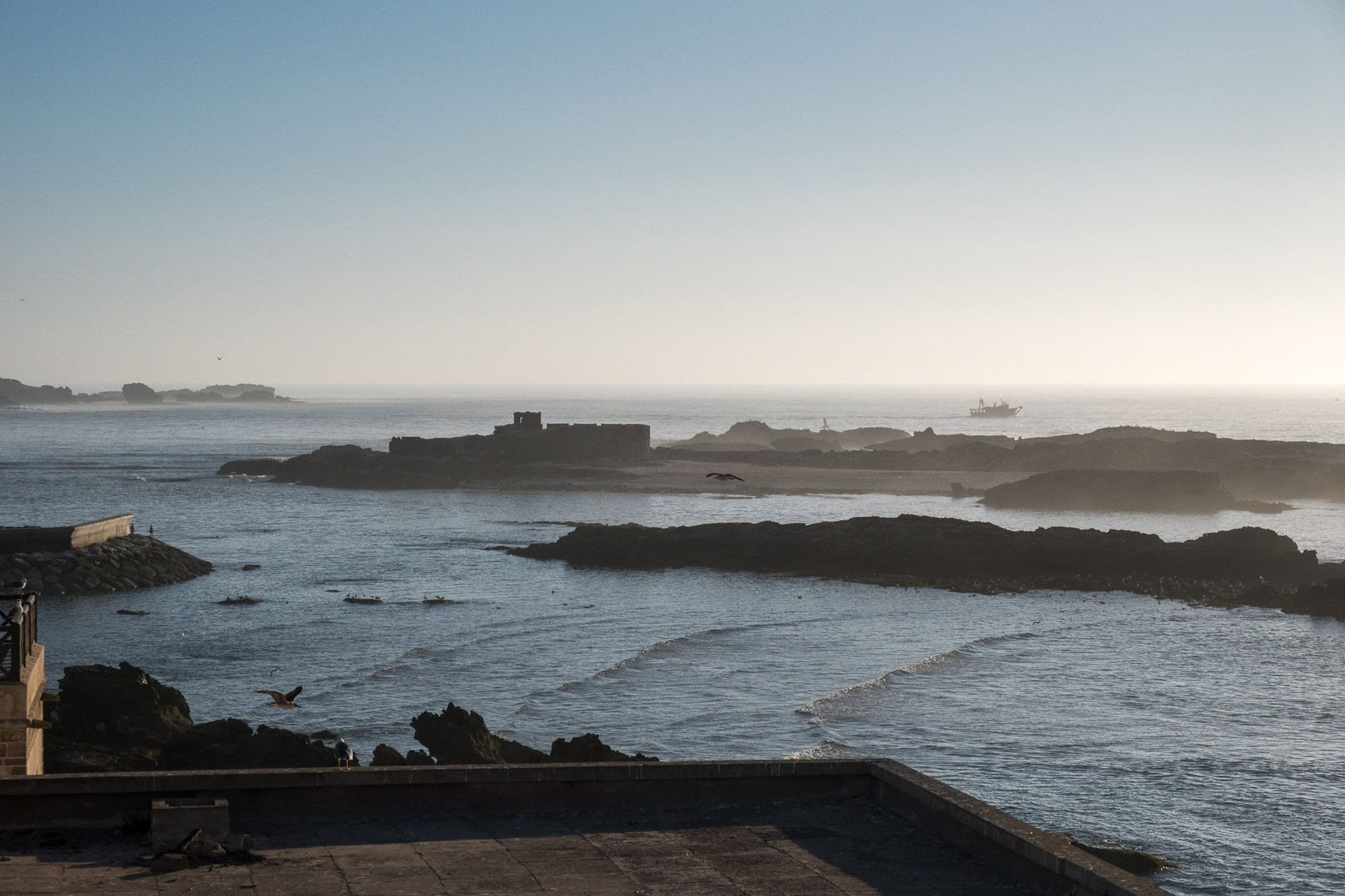 Coastal scene at sunset with rocky shoreline, calm sea, distant boat, and hazy sky.
