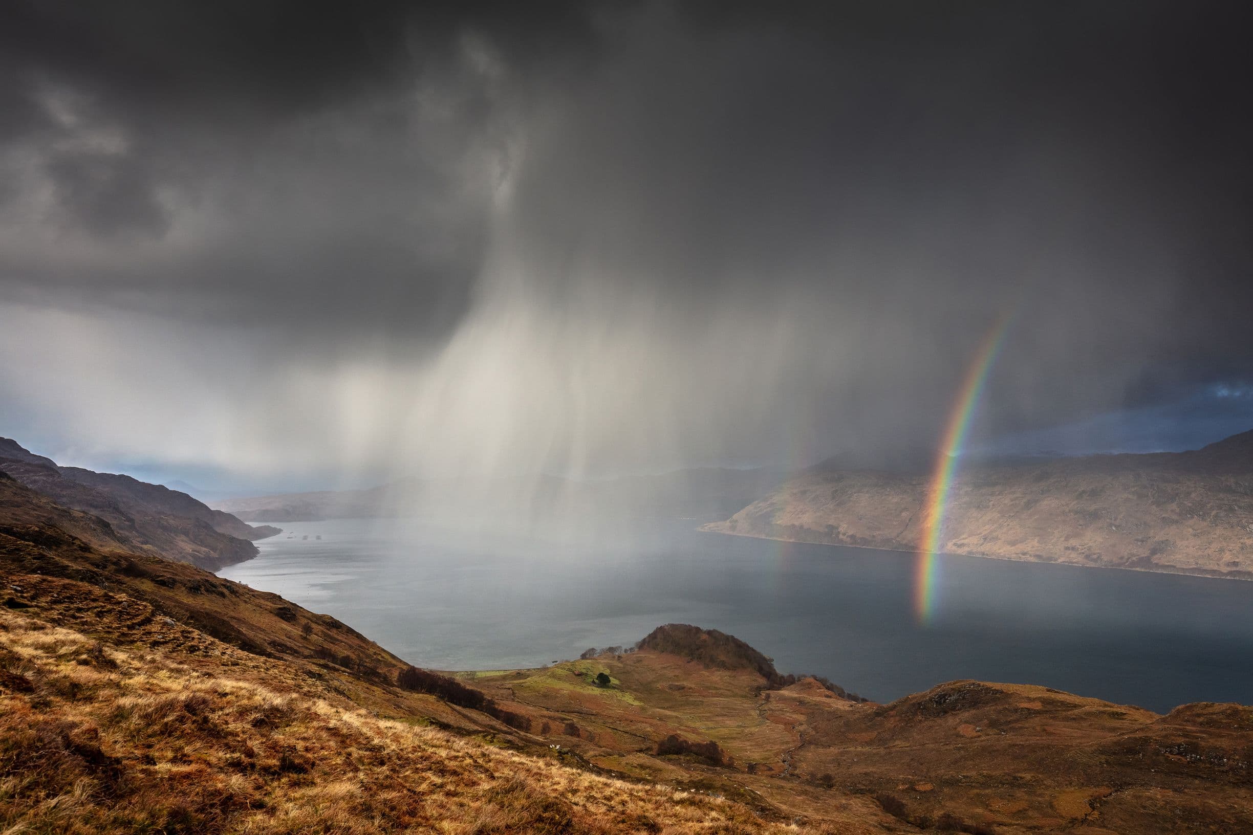 A dramatic landscape with dark storm clouds, rain over a lake, and a vibrant rainbow arching across the scene.