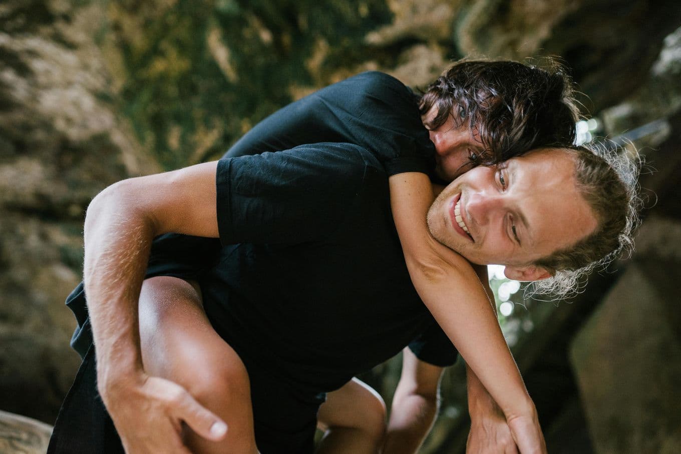 A person gives another a piggyback ride in a natural setting, both smiling and wearing black shirts, with blurred greenery in the background.