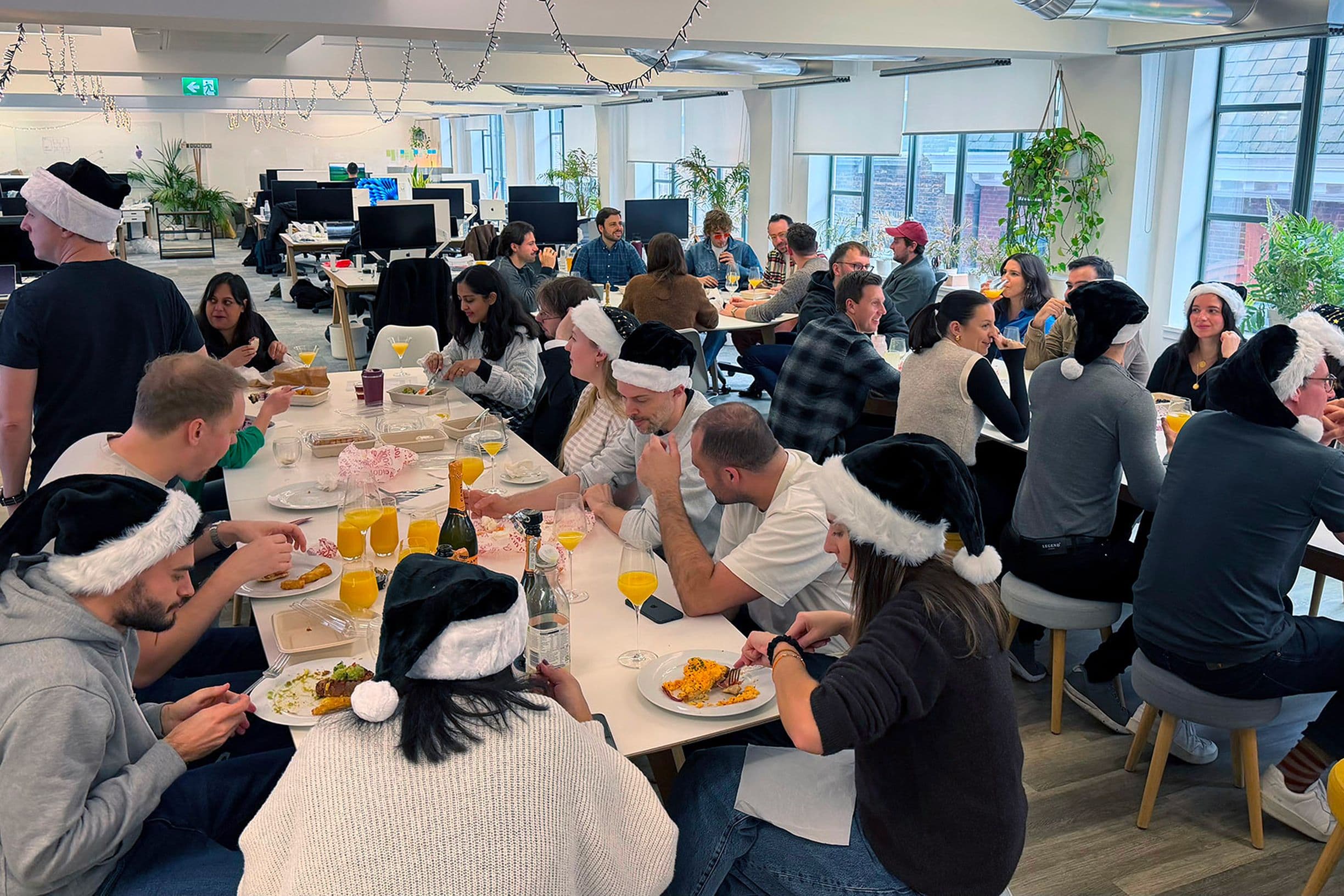 A large group of people wearing Santa hats, sitting and eating together in a bright office decorated for the holidays.