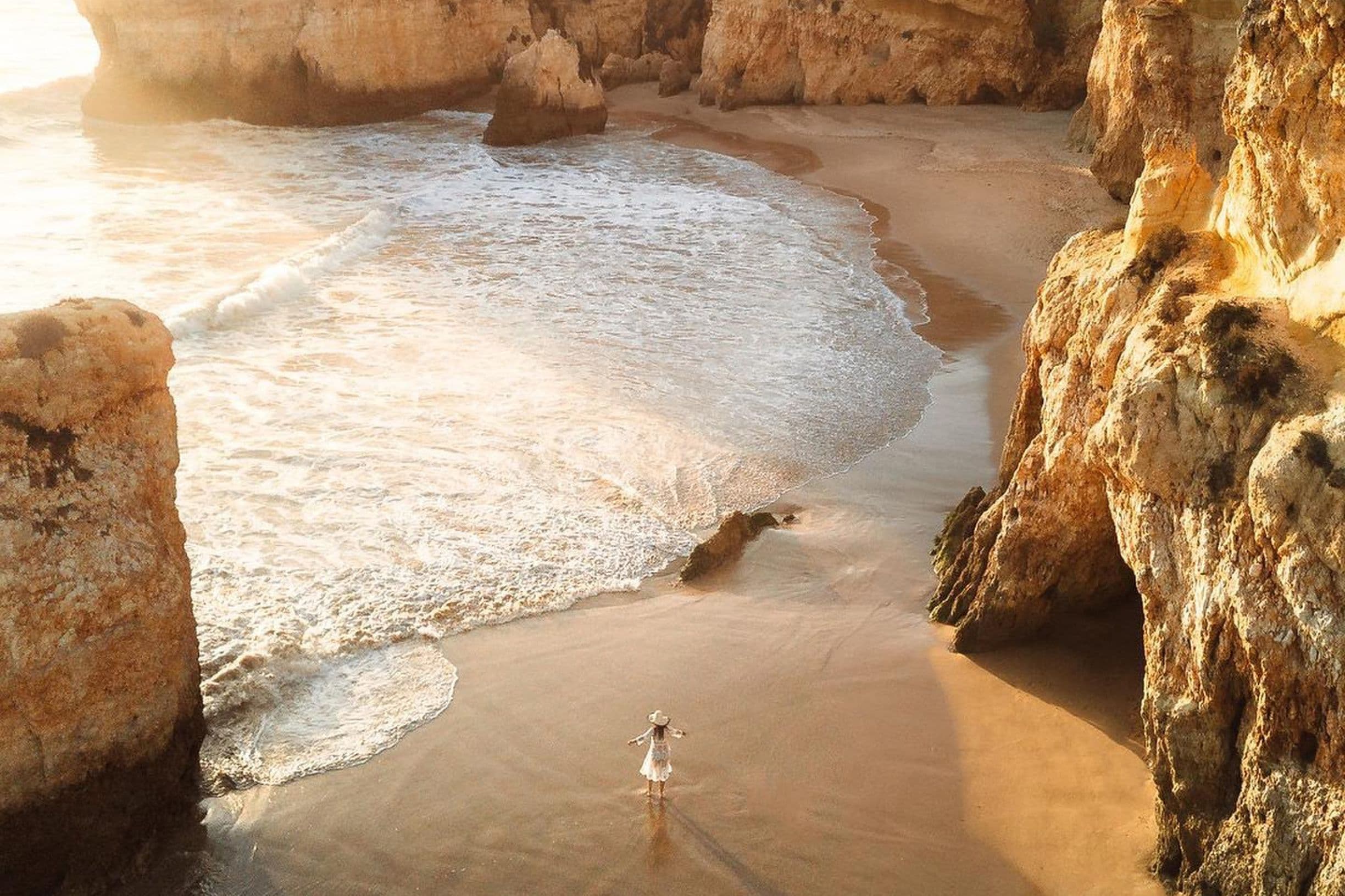 Person in a white dress stands on a sandy beach surrounded by cliffs, with gentle waves lapping at the shore during sunset.