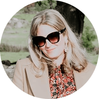 Woman with long hair wearing sunglasses, a floral top, and a beige coat, smiling outdoors in a sunny setting.