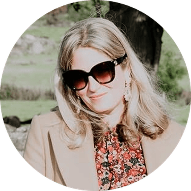 Woman with long hair wearing sunglasses, a floral top, and a beige coat, smiling outdoors in a sunny setting.