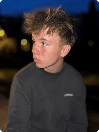 Teen with tousled hair and a black shirt gazes to the side against a blurred, dimly lit background.