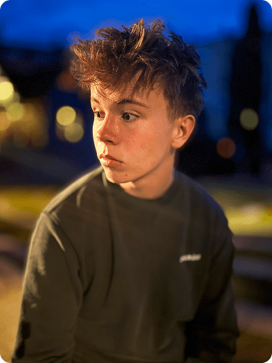 Teen boy with tousled hair looks to the side, wearing a dark shirt, with a blurred evening background.