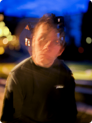 A person with a blurred face and double exposure effect, sitting outdoors at night with bright bokeh lights in the background.
