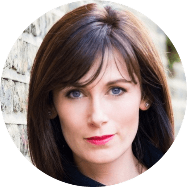 Close headshot of a woman with shoulder-length brown hair, blue eyes, fair skin and pink lipstick, posed against a brick wall.