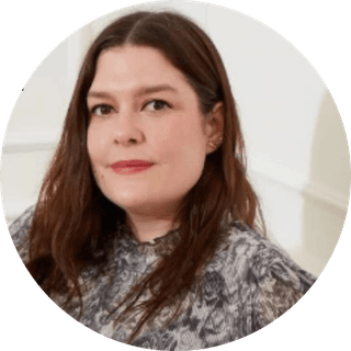 A woman with long brown hair wearing a patterned blouse, standing against a plain white background.