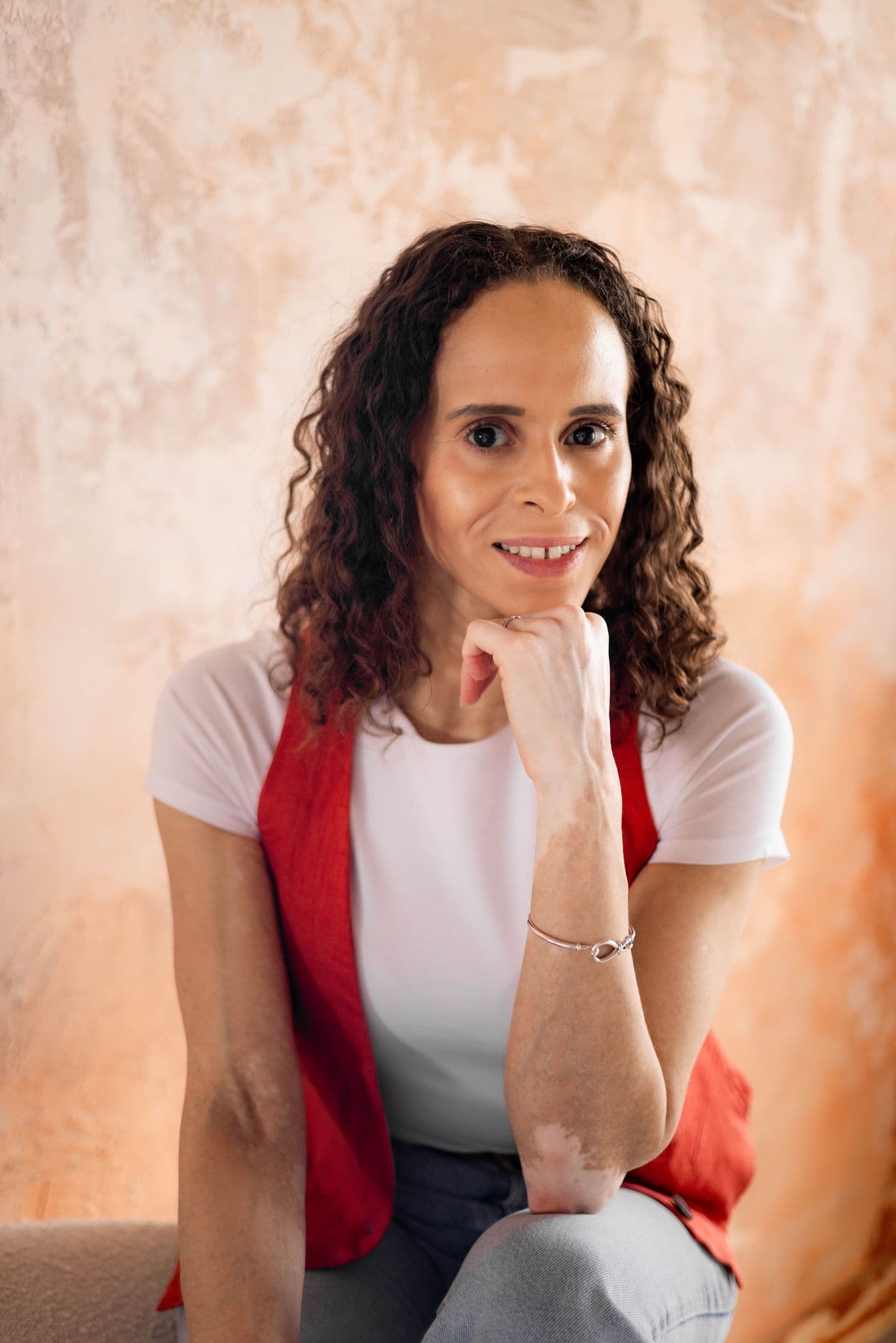 Natalie Ambersley wearing a red vest and white shirt smiles, resting their chin on their hand against a textured, peach-coloured background