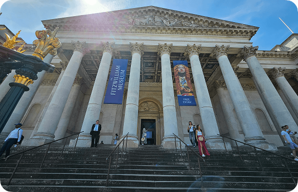 Front view of the Fitzwilliam Museum with neoclassical columns, banners, and people on the steps under a clear blue sky.