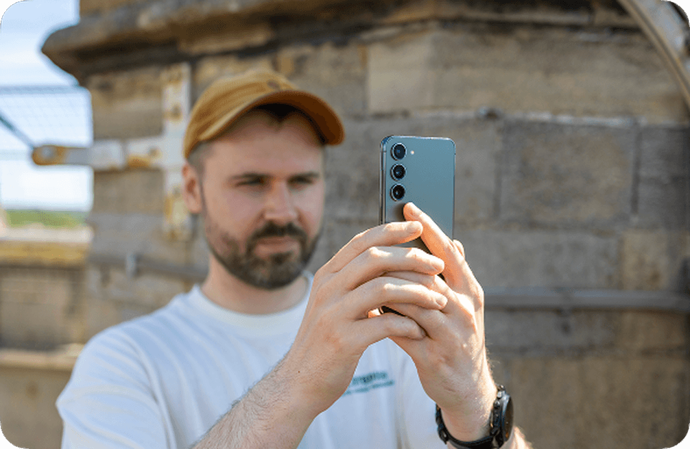 Dan, wearing a cap, takes a photo with a smartphone outdoors, standing near a stone structure.