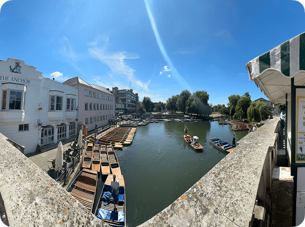 A scenic riverside view with multiple boats docked, buildings lining the water, and a clear blue sky overhead.
