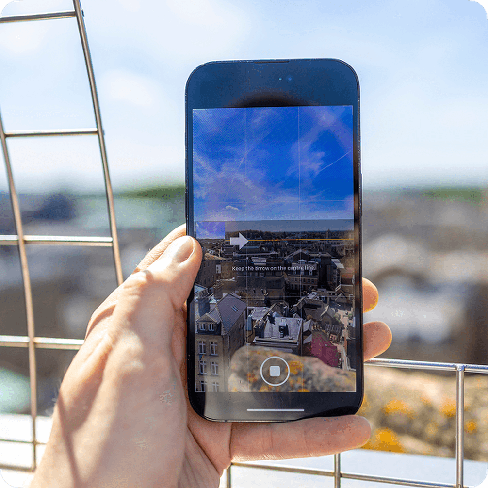 Hand holding an iPhone in panorama mode capturing a panoramic cityscape from a rooftop, with a metal fence in the background.