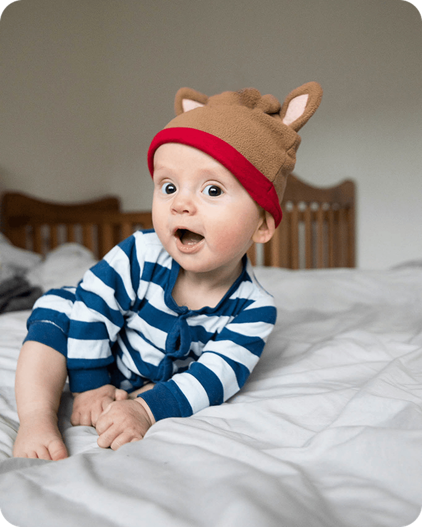Baby in blue striped pajamas and a brown hat with ears, sitting on a bed, looking surprised.