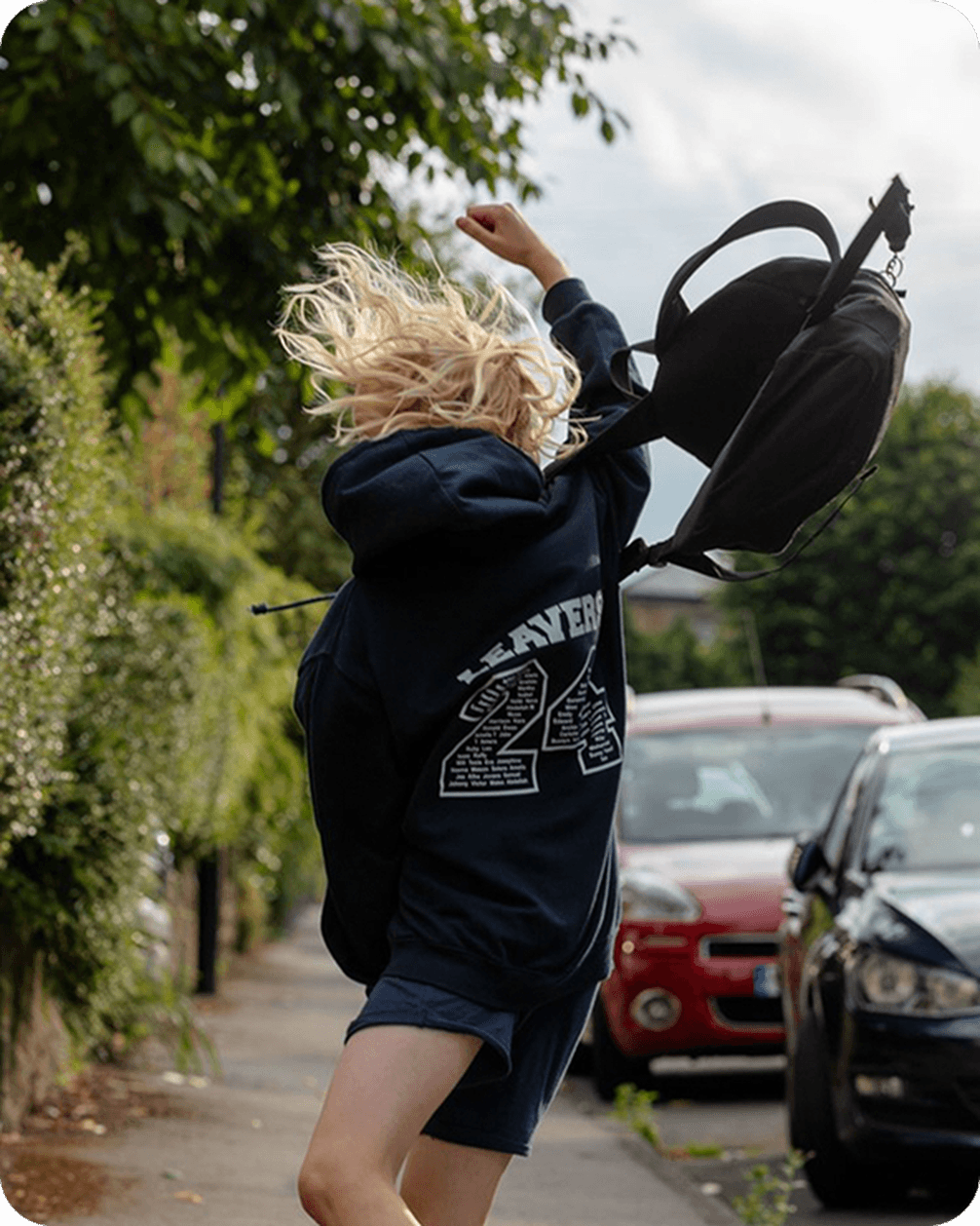 Person in a hoodie joyfully jumps on a sidewalk, with a backpack in the air. Cars and greenery line the street.