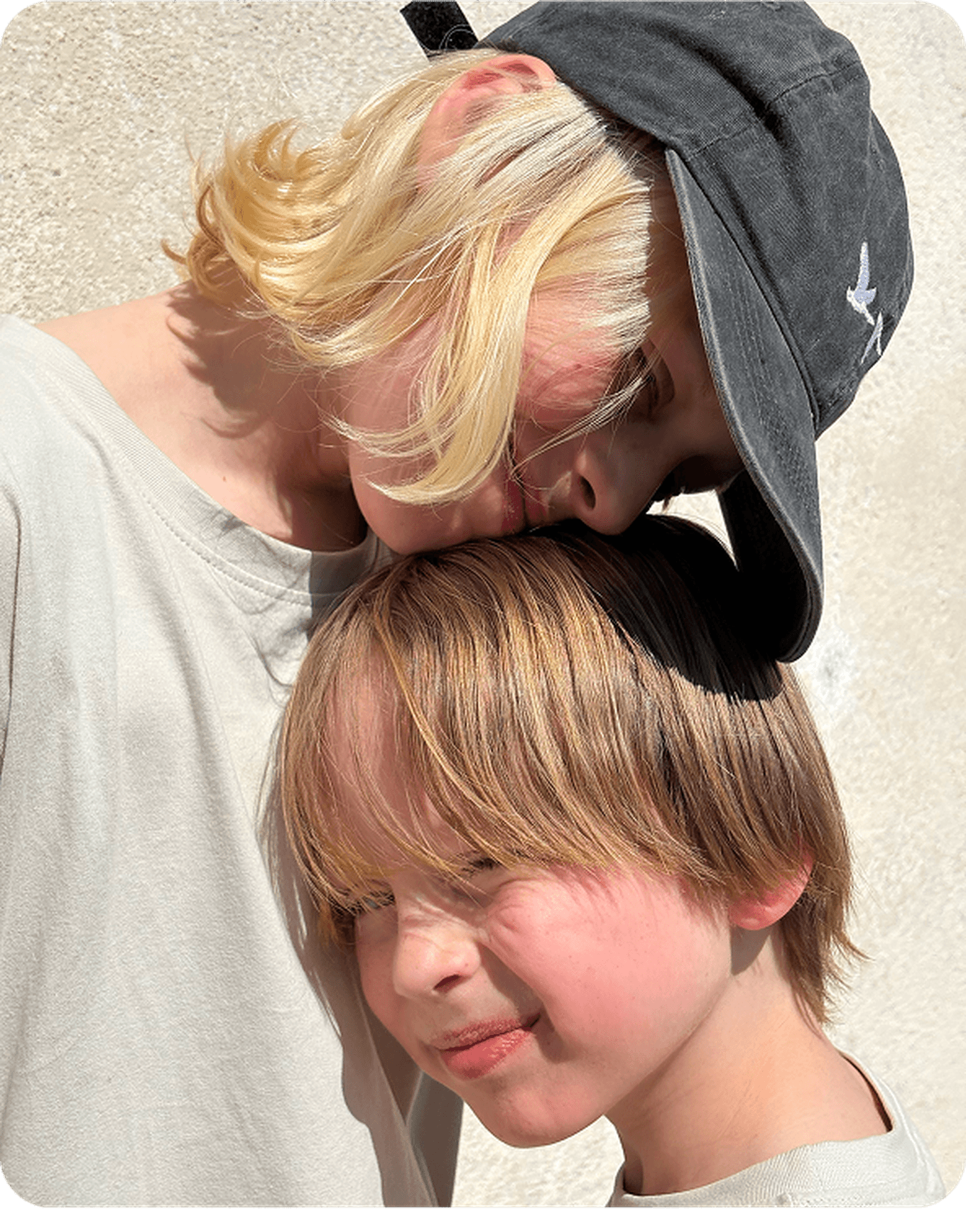 Two children with light hair share a tender moment. One wears a cap and rests their head on the other's, both smiling softly against a light background.