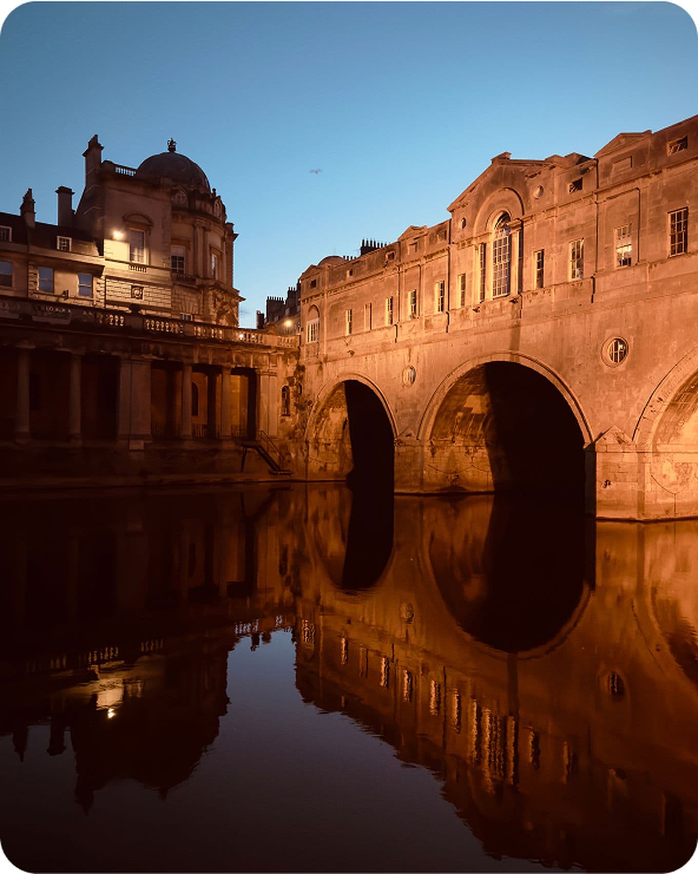 Historic stone bridge and buildings reflected in calm water at dusk, under a clear sky with warm lighting.