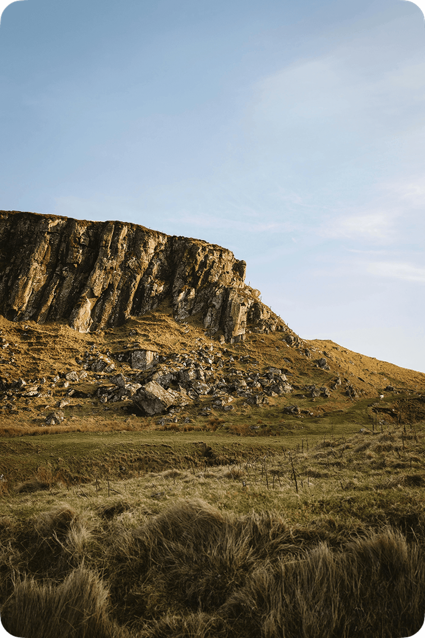 A rugged cliff rises above grassy terrain under a clear blue sky, with scattered rocks and a gentle slope.