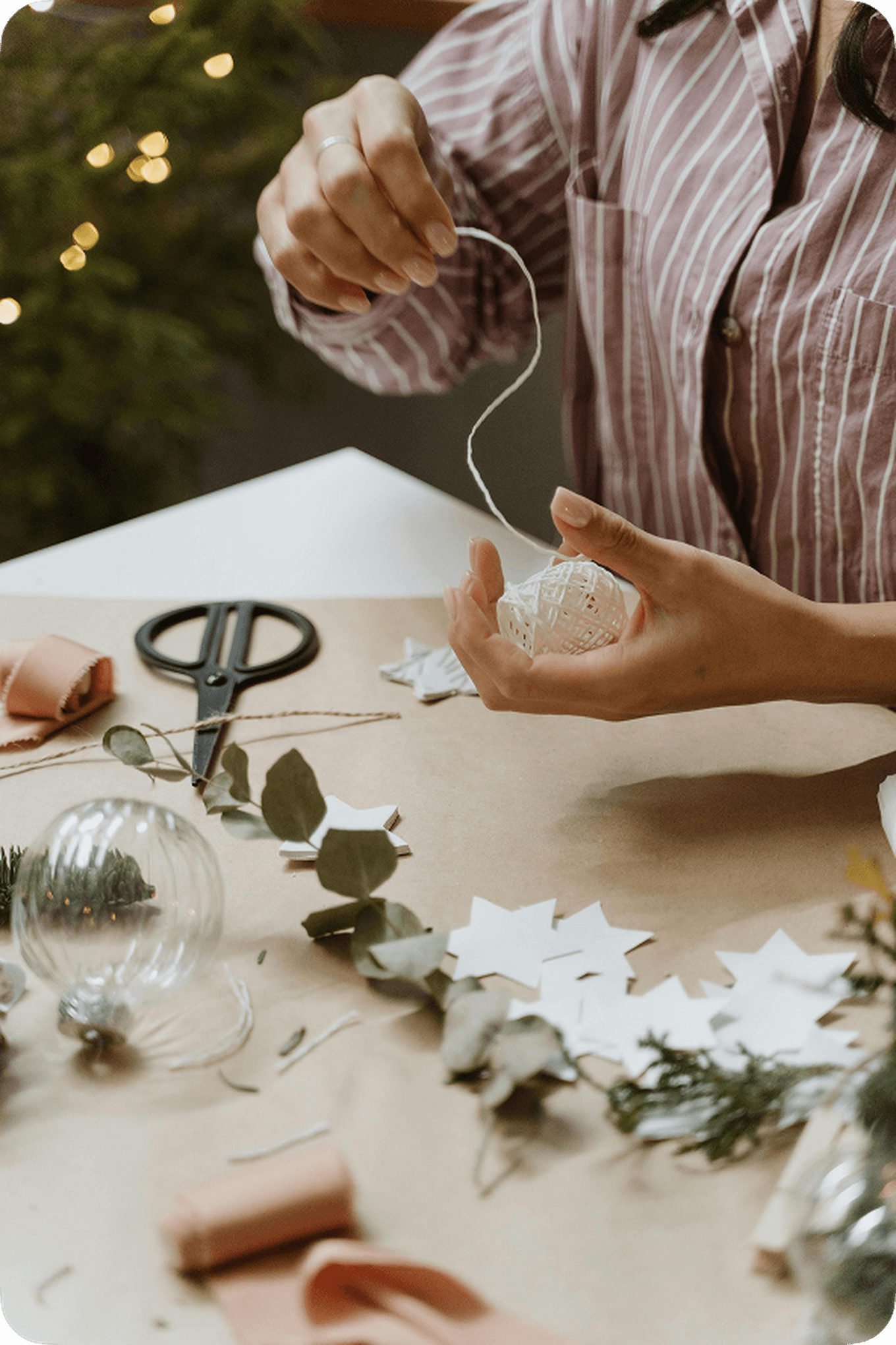 Person in striped shirt crafting with twine and clear ornament, surrounded by paper stars, greenery, scissors, and ribbon on a table.