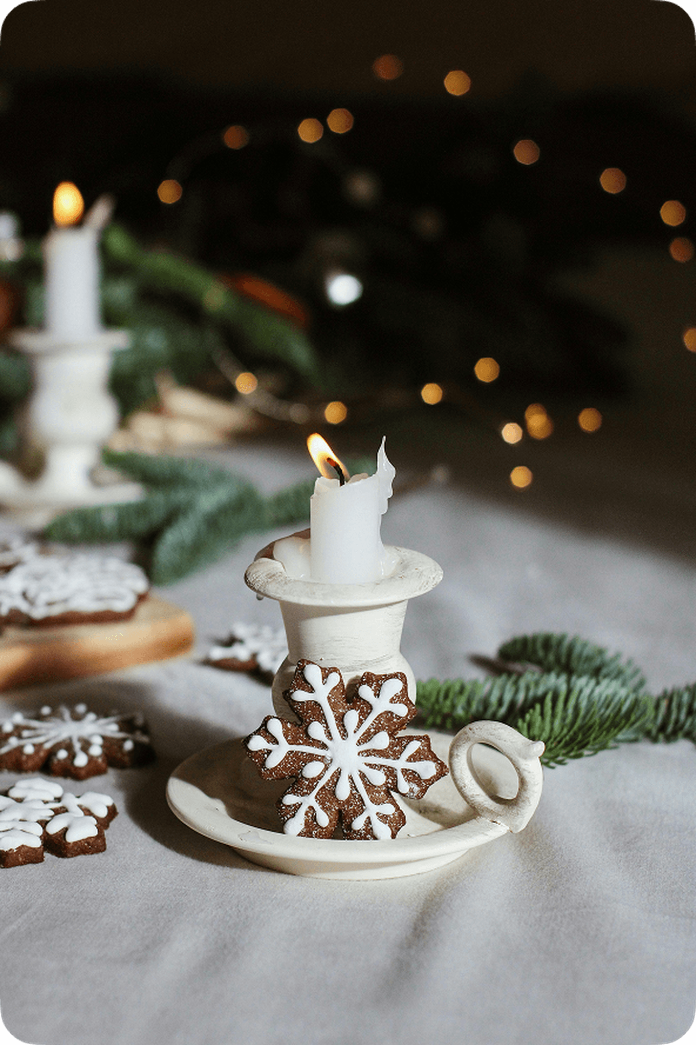 Lit candle in a holder with a snowflake cookie, surrounded by greenery and soft lights on a table.