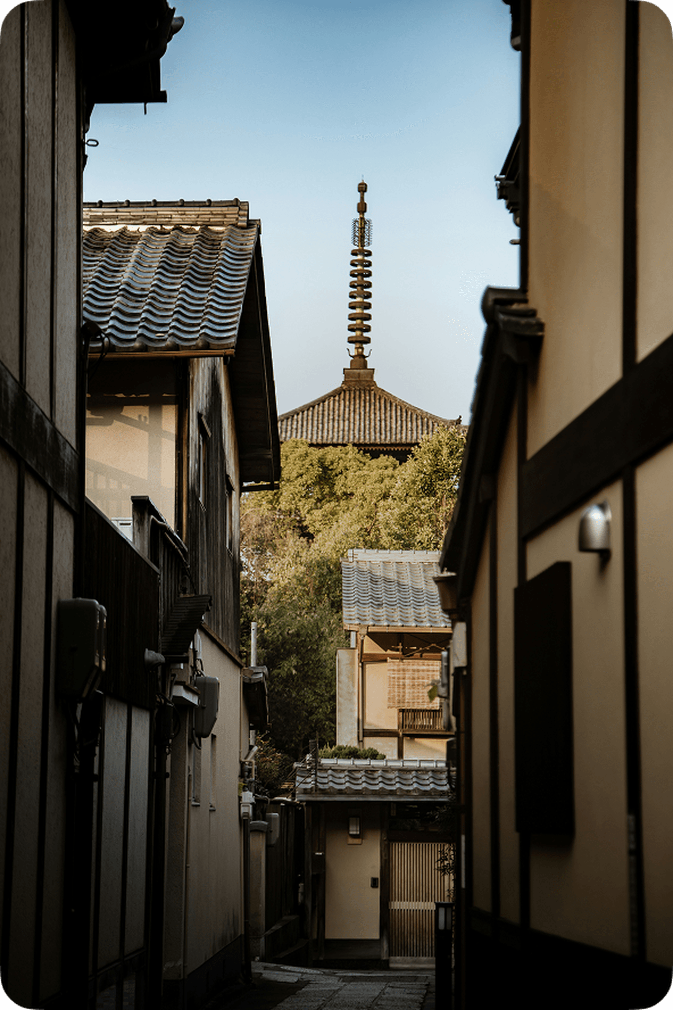Narrow alley with traditional Japanese buildings, leading to a view of a pagoda surrounded by trees under a clear sky.