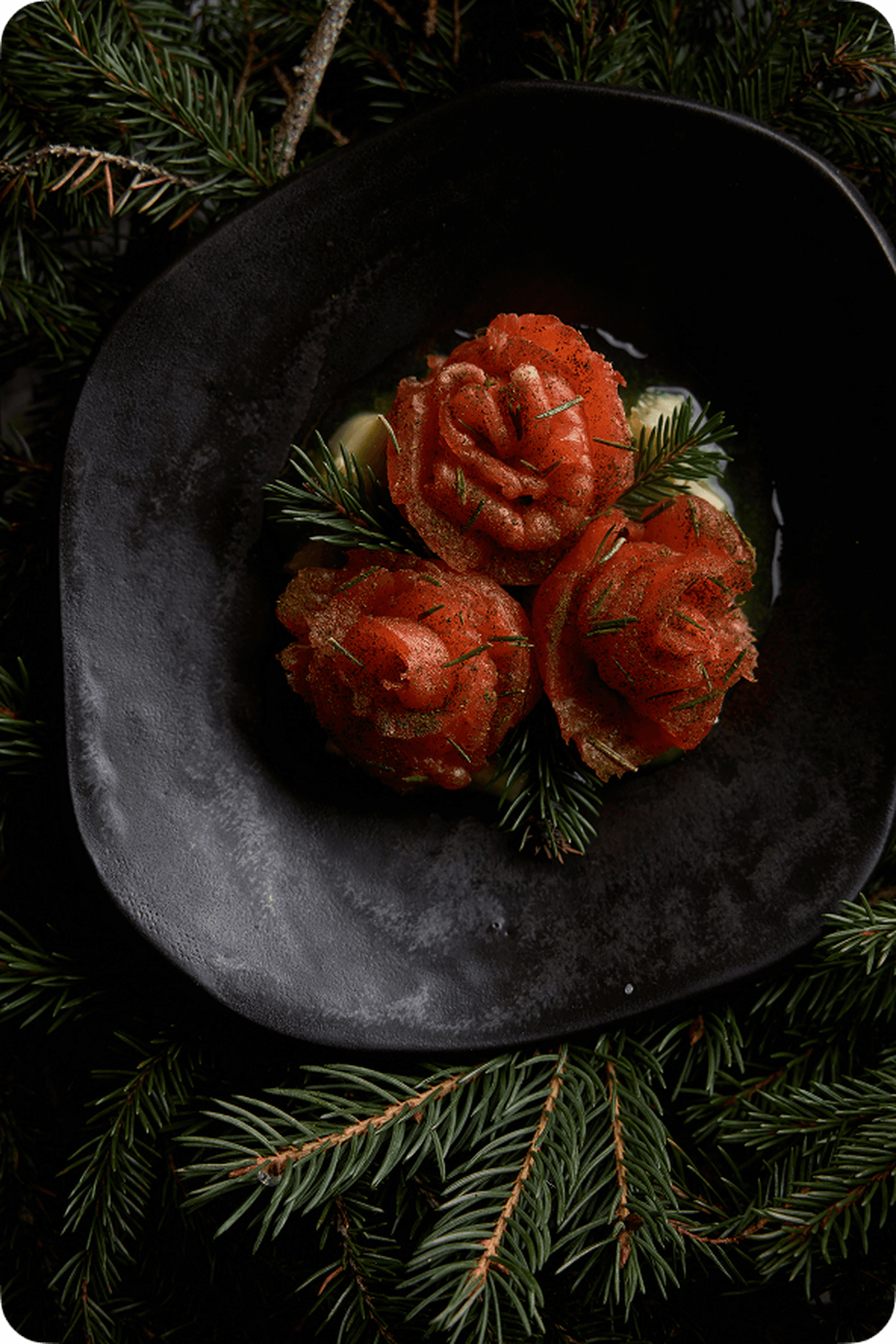 Three salmon rosettes garnished with dill on a black plate, surrounded by pine branches.
