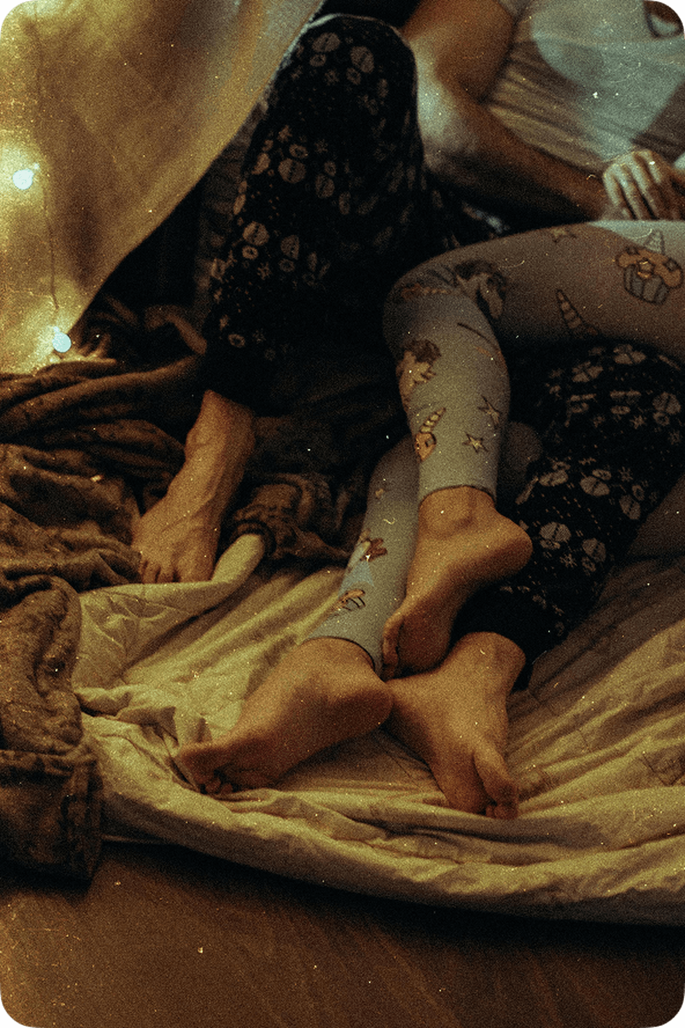 Two people in cosy pajamas, legs intertwined on a bed with soft blankets and warm string lights in the background.