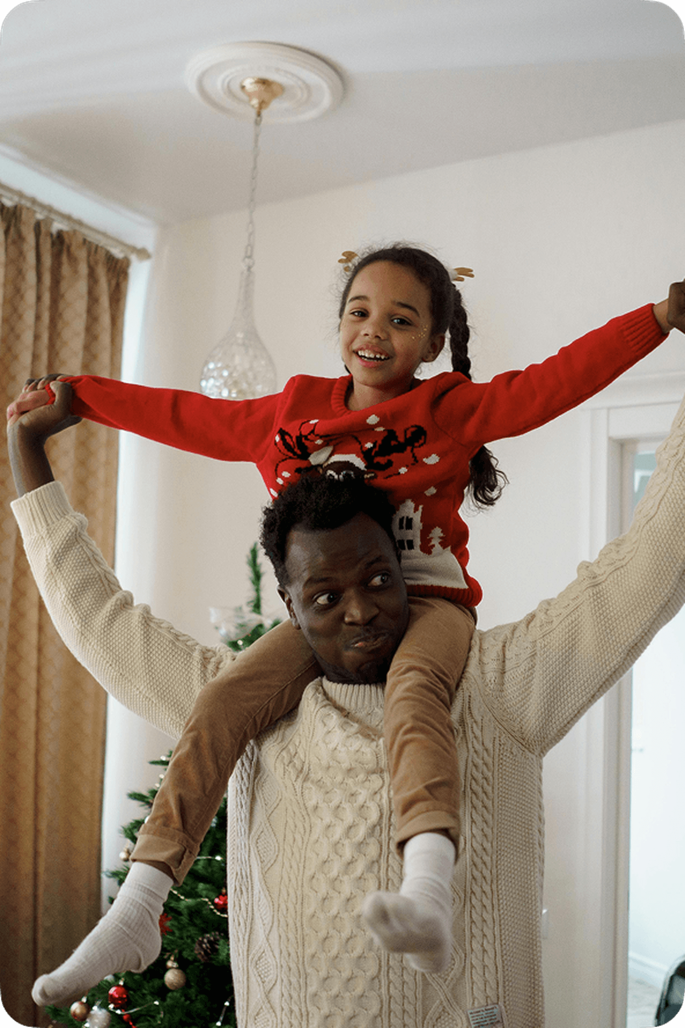 Man in a white sweater holding a smiling child on his shoulders. The child is wearing a red sweater. A decorated Christmas tree is in the background.